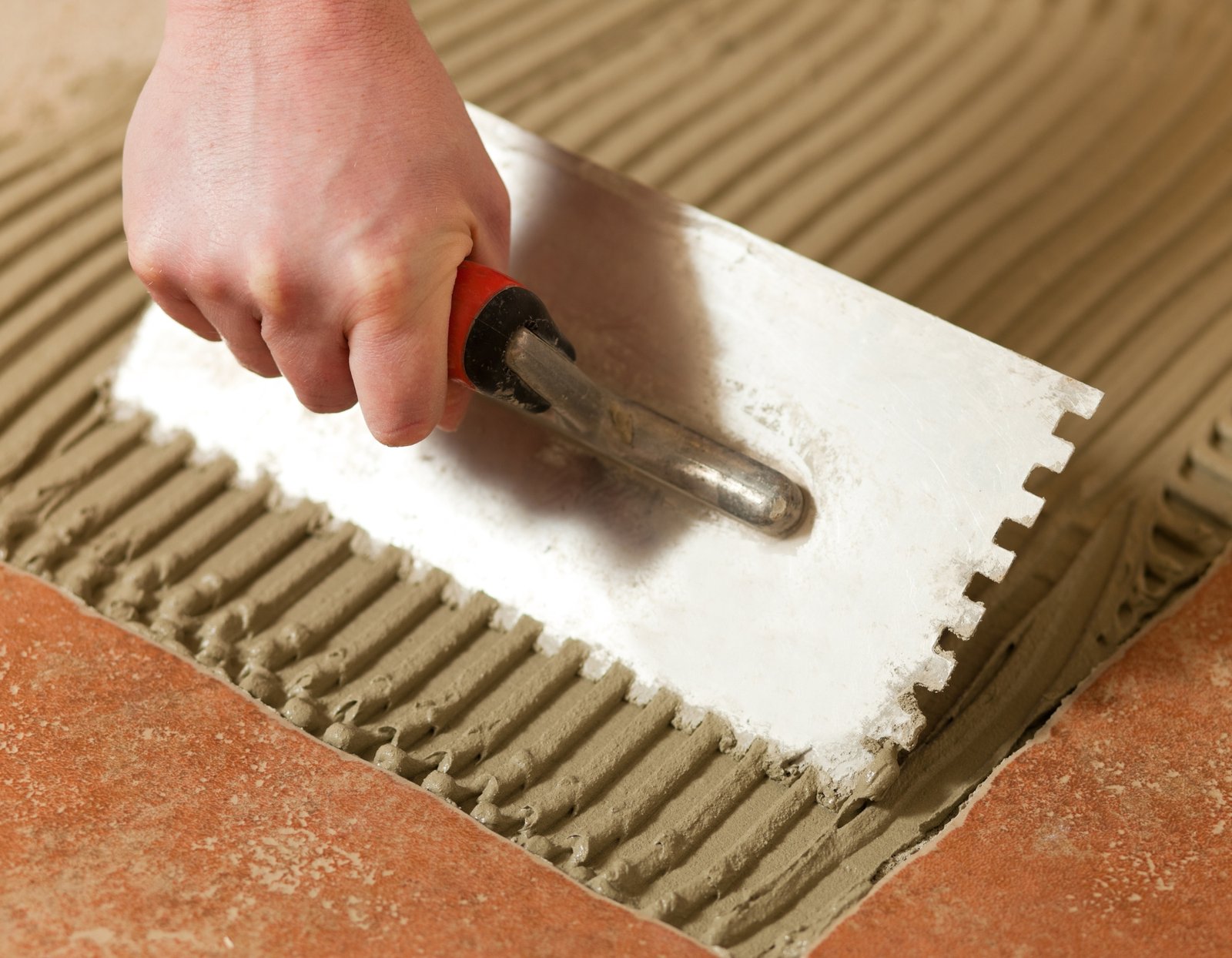 Een hand brengt vloerlijm aan met een troffel tijdens tegelwerken.